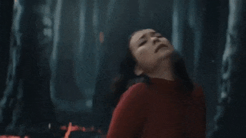 Music video gif. Mitski is in a fiery forest and looks as if she's in pain. She throws a hand out and moves towards us jerkily, as if she's being summoned and cannot resist.