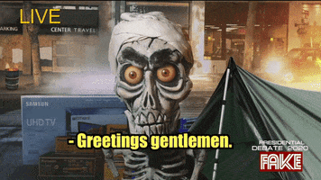 Video gif. Jeff Dunham's Achmed the Dead Journalist, a skeleton puppet, reports from a burning city, a tent and a UHD TV box behind him, raising his eyebrows as he talks to us, saying, "Greetings gentlemen."  