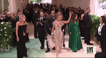 Met Gala 2024 gif. Jennifer Lopez waves and blows a kiss at fans and press. She's wearing a near-see-through Schiaparelli sleeveless gown adorned with stone embellishments, fitted through the waist with a short train and  detailing at her shoulders that resembles peacock feathers. She has neutral makeup and her hair is swept back in a high bun. 