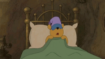 Disney gif. Winnie the Pooh hugs his pillow and snuggles in bed while wearing a sleeping eye mask and night cap.