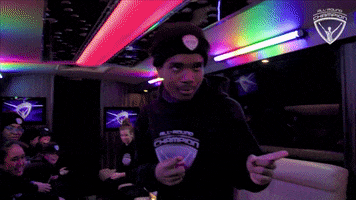 Video gif. Beneath multicolored lights on a low ceiling, a smiling young man makes finger guns to right of frame.