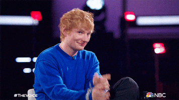 Reality TV gif. Ed Sheeran as a guest on The Voice, points and smiles at someone off screen with both hands.