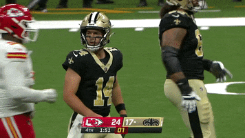 Sports gif. Jake Haener from the New Orleans Saints pauses on the field, holding a hand up to his ear like, "I can't hear you." Players run in all directions behind him as a teammate slaps him on the helmet. 