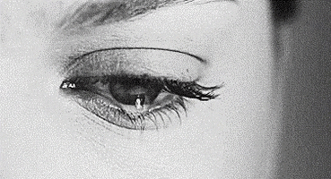 Video gif. Black and white close-up of an eye with long lashes and a tear slipping out.
