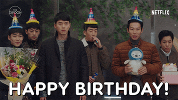 Movie gif. Hyun Bin as Ri Jeong-hyeok in "Crash Landing on You" and a group of friends all stand in a line wearing colorful birthday hats and holding gifts, including a bouquet of flowers, a cake, and a stuffed animal. Bin stands still, looking off expectantly like he's waiting for someone.