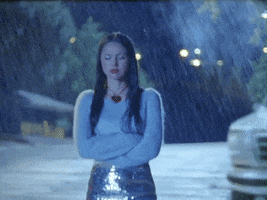 TV gif. Olivia Rodrigo walks down a suburban street in the pouring rain with her arms crossed, looking from side to side with an angry and annoyed expression, dropping her head with a sulking look.