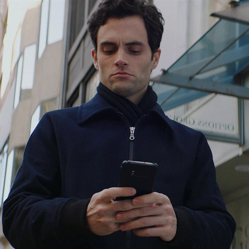 TV gif. Penn Badgley as Joe on the show "You" stands outside of a building and looking with concern at his phone.