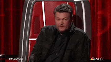 Reality TV gif. Blake Shelton as a judge on The Voice Turns to someone off screen, raising his hand in a gesture of questioning. 