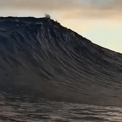 Video gif. An ocean wave, gathers, then crests, in slow motion.