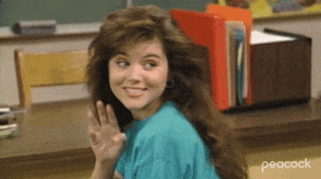 TV gif. Tiffani Thiessen as Kelly Kapowski on Saved By The Bell looks over her shoulder as she sits at her desk in school. She has a flirtatious smile and she wiggles her fingers to playful say hello. 