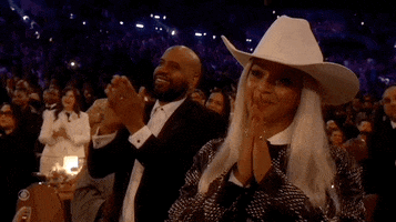 Celebrity gif. Beyonce wears a white cowboy hat and a black-and-white checkered jacket as she claps enthusiastically at the Grammy Awards.
