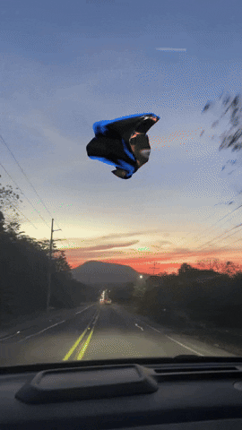 Digital art gif. From the driver's seat of a moving car, we see an amorphous latex blob floating midair and reflecting off bold, neon colors against a dusky sky. 
