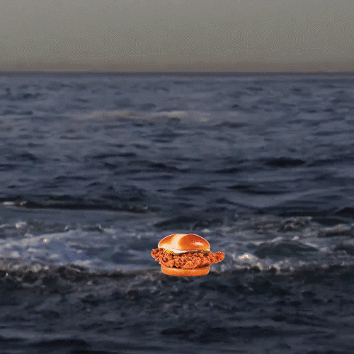 Video gif. A Great White Shark breaches the water and snaps up a Bojangles Chicken Sandwich in its mouth.