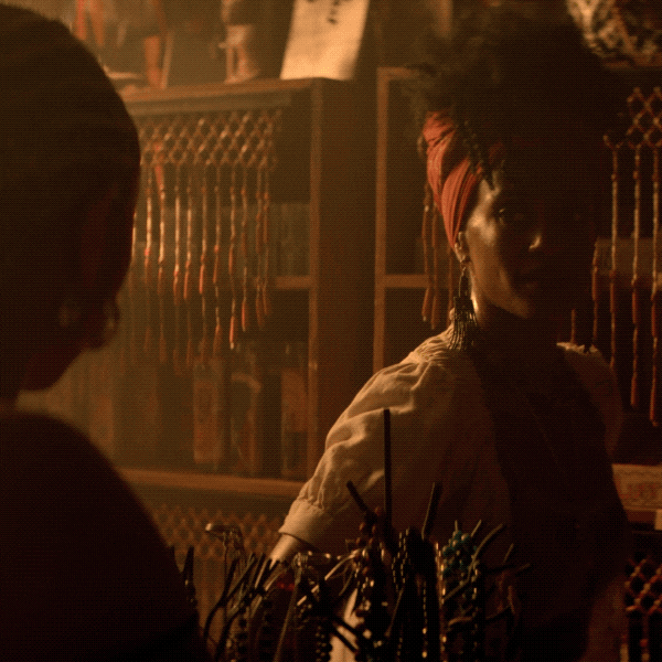 TV gif. Tati Gabrielle as Prudence looks at Skye P. Marshall as Marie LaFleur in "The Chilling Adventures of Sabrina." The two are in a voodoo shop wearing Haitian-inspired clothes. Gabrielle smiles slightly, a feeling of mischief in the air.