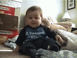 Video gif. Low-angle footage of a giggling baby sitting on a living room floor. As the baby continues laughing, he gently topples over to one side and rolls onto his stomach.