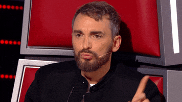 Reality TV gif. Christophe Willem on The Voice Belgium sits in the red chair, pursing her lips and wagging his finger side to side.