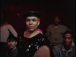 Movie gif. Drag performer Paris Dupree in Paris is Burning glimmers in a sequined black top and a black beret, soaking up the spotlight as she wraps her leg tightly around a man's neck. She grabs onto her foot with one arm and vogues with the other while the man lets out choked laughter into her arm.