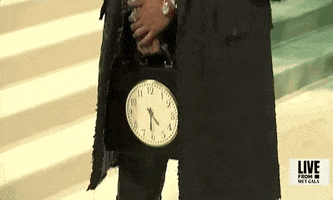 Met Gala 2024 gif. Camera pans up on Ben Simmons holding a black rectangular briefcase with a clock on the front. He wears a black leather overcoat that hangs over his shoulders and circular rose-colored sunglasses. 