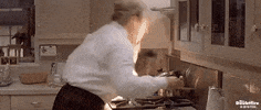Movie gif. Leaning over a burner, Robin Williams as Mrs. Doubtfire freaks out when her blouse catches fire and tries to tamp it out by pounding a spatula on her chest.