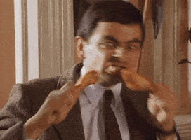 TV gif. Rowan Atkinson as Mr. Bean takes fast, alternating bites from two chicken drumsticks.