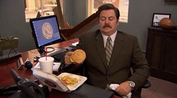 Parks and Recreation gif. Nick Offerman as Ron at his desk with a takeout container, holds a burger and then chucks it at his open mouth, where it bounces off.