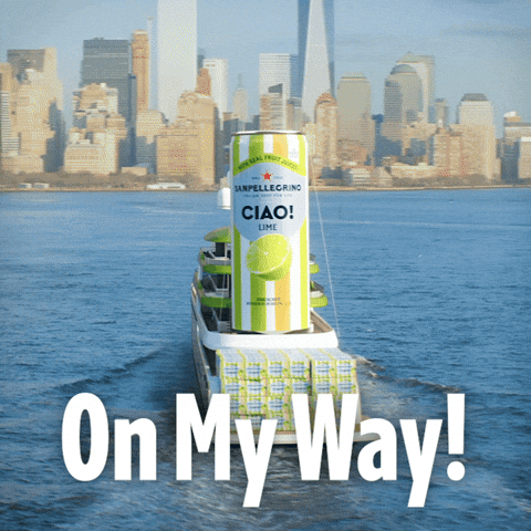 Sponsored gif. Ferry boat heads toward New York City. The flag of Italy flies from the top of it and the body of the ferry is covered in green, white, and orange stripes. On the back of the ferry are stacks of cases filled with Sanpellegrino. Above the boxes is a massive striped can of Sanpellegrino Ciao! Lime. Text reads, “On my way!”