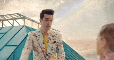 Music Video gif. Brendon Urie in his music video with Taylor Swift for their song called ME! He gives Taylor a big bundle of bright flowers and looks anticipatory at her response.