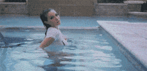Video gif. Woman slowly stands up in a pool with a soaking well shirt clinging to her body. She looks at us with a sexy, flirtatious expression.