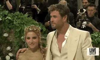 Met Gala 2024 gif. Close on Chris Hemsworth and Elsa Pataky as they for the cameras in coordinated Tom Ford ensembles, zoom out to show Chris's sand-colored suit, and Elsa's clingy golden turtleneck gown.
