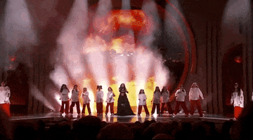 Oscars 2024 GIF. Wide shot of Becky G and a girls' choir performing The Fire Inside from Flamin' Hot. Becky G and the girls rock side to side and the girls are all wearing a white top and red pants. Flames shoot up behind them.  