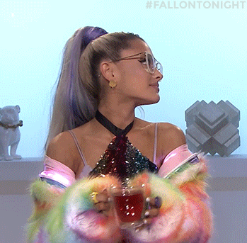 TV gif. Ariana Grande on The Tonight Show wears fluffy rainbow sleeves as she smiles and takes a sip of tea. 