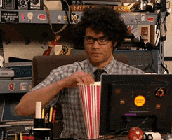 TV gif. Richard Ayoade as Maurice in The IT Crowd sits at his desk and looks smugly at someone off screen as he eats a piece of popcorn. 
