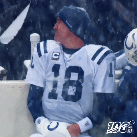 Sports gif. Payton Manning of the Colts sits on the sidelines as snow falls down heavily around him. He looks over at someone and gives a thumbs up.