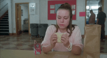 Movie gif. Tina Majorino as Deb in Napoleon Dynamite. She awkwardly sits at a lunch table and eats a sandwich with a juice box next to her. She's clearly uncomfortable as she looks down and squirms in her seat while eating.