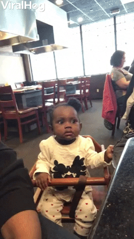 Video gif. A baby girl at a restaurant, sitting in a high chair. Her eyes grow large as her face is lit up by the flame from a hibachi grill. She looks side to side, before reaching out to the person next to her.