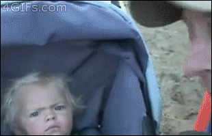 Video gif. A blonde baby in a stroller looking over at someone with suspicion.