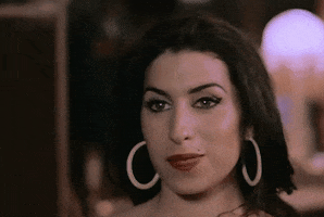 Music video gif. Amy Winehouse in her video for In My Bed, with a shy smile, waves at us flirtatiously.