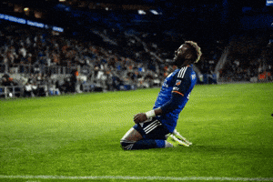 Sports gif. Aaron Boupendza of FC Cincinnati slides across the soccer field on his knees in celebration after scoring a goal and his head is tossed back in a triumphant roar as he clenches his fists. His teammates surround him and grab his back, celebrating together.
