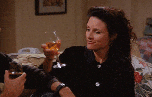 Seinfeld gif. Julia Louis-Dreyfus as Elaine clinks wine glasses with a man and smiles eagerly and dorky, biting her lower lip.