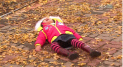 Video gif. On a brick walkway with fallen leaves scattered around, a child in a red coat lies on her back and flails around in a tantrum.