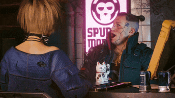 Video game gif. We zoom in over and over on Jackie Welles from "Cyberpunk 2077" seated across a desk from another person as he turns to look at us with a surprised expression. He has geometric face tattoos and wears a black tactical jacket with a high collar. 