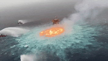 Video gif. Oil fire in the ocean off the Gulf of Mexico. Three ships are spraying water on it, trying to put it out, but the circle of fire rages on.