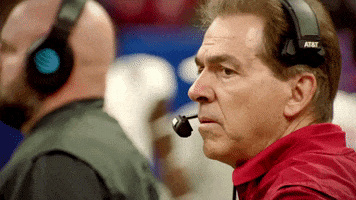 Sports gif. Coach Nick Saban stands on the sidelines with headphones on his head. He looks over with a worried expression on his face.