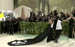 Met Gala 2024 gif. Laurie Cumbo and Bobby Digi Olisa ascend the stairs in matching black-and-white ensembles, an assistant holding her painted train.