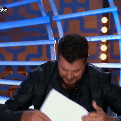TV gif. Luke Bryan as a judge on American Idol fans himself with his papers as he's gotten hot from seeing a contestant's talent.