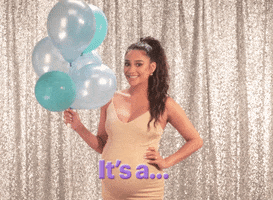 Celebrity gif. Shay Mitchell, holding gray and blue balloons, rubs her hand over her pregnant belly saying, “It’s a boy!”