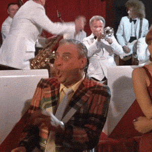 Movie gif. Rodney Dangerfield in Caddyshack, an elderly white man in a loud plaid suit, dances obnoxiously to a live band among elegant partygoers. He dips and sways his hips, snapping his fingers erratically and making cartoonish facial expressions. 