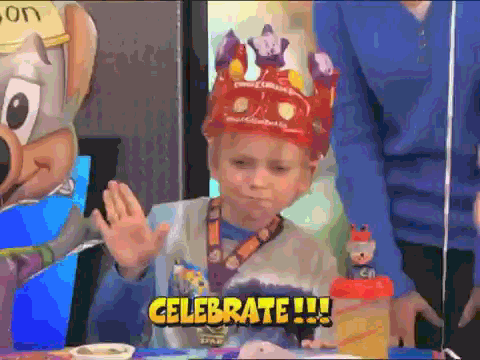Malý tancující chlapec s korunou, stojící před stolem s dobrotami a nápisem "Celebrate!". 