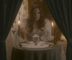 Video gif. Woman dressed as a fortune teller sits at a circular table with candles and tarot cards. She waves a hand over a crystal ball and text appears, "It's giving... Bleak..." She looks up and gives us an insincere concerned expression. 