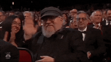 Celebrity gif. George R.R. Martin sitting in the audience at the Emmy Awards waving at us.